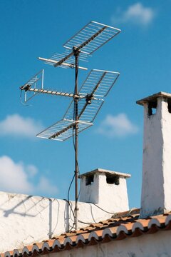 Old style TV antenna sits on rooftop under clear blue sky. Chimneys and tiled roof suggest older residential building. Antenna receives broadcast signals for home viewing entertainment.