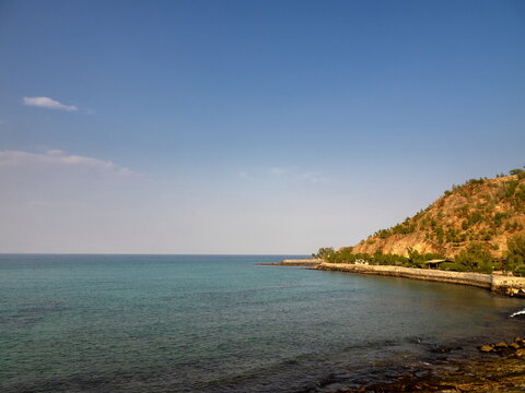 View of the turquoise sea meeting the rocky coastline with sparse vegetation on the hill under a clear blue sky in Ras Al-Khaimah, Ras Al Khaimah, United Arab Emirates.