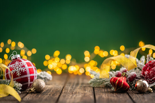 A Christmas background featuring a red plaid bauble with a snowflake and gold ribbon, red ornaments, and frosted berries on rustic wood, set against green with twinkling lights and copy space.