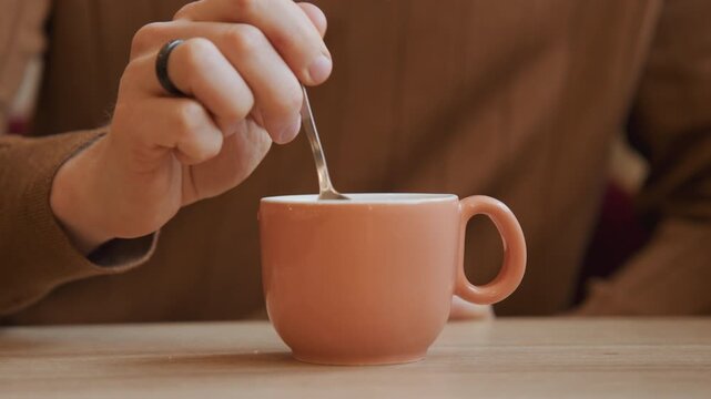 Close up of hand stirring liquid in pink ceramic cup on wooden table. person wearing brown sweater and ring repeatedly stirs drink with spoon throughout sequence. Soft lighting creates warm