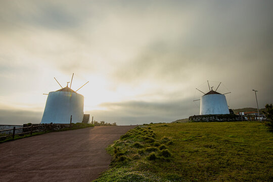 Corvo, Azores, Portugal Two white windmills stand tall on grassy hillside at dusk. Pathway winds toward them under cloudy sky. Warm golden light spills behind structures. Scene feels calm and nostalgi