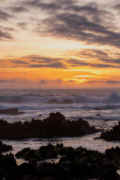 Corvo, Azores, Portugal Sunset paints sky orange and purple over rocky shore. Waves crash against dark volcanic rocks. Sky clouds glow warm with fading daylight. Sea mist rises near waterline. Mood is