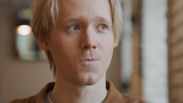 Close up of man with short blonde hair wearing brown shirt indoors. Man holds and inserts acupuncture needle into his lower lip demonstrating process in detail against softly lit background