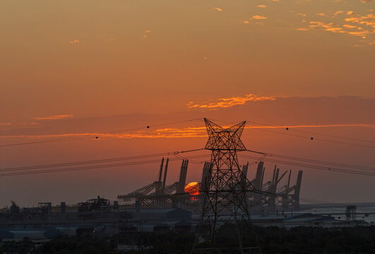 View of a large electrical transmission pylon silhouetted against a vibrant sunset sky with industrial port cranes in the background in Dubai, Dubai, United Arab Emirates.