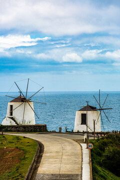 Corvo, Azores, Portugal Two white windmills stand on a cliff overlooking the blue ocean. Winding path leads toward them. Clouds drift above the sea and mill structures. The scene is peaceful and sceni
