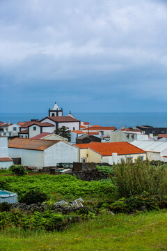 Corvo, Azores, Portugal Coastal village with white houses and red-tiled roofs. Church spire rises above clustered homes near ocean. Green vegetation covers foreground, adding contrast to warm tones. S