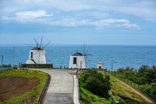 Corvo, Azores, Portugal Two white windmills stand on a coastal hill, overlooking blue sea. Sky is partly cloudy, casting soft light on scene. Path leads toward mills, inviting viewers to explore. Gree