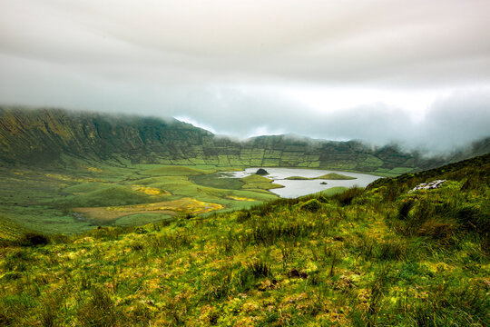 Corvo, Azores, Portugal Lush green hills roll toward misty valley. Small lake reflects cloudy sky above. Wildflowers dot foreground grasslands. Mountains fade in soft fog layers. Scene feels calm, qui