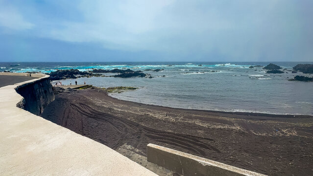 Corvo, Azores, Portugal Coastal view with dark volcanic sand and calm waters. Concrete walkway curves along shore under bright sky. Horizon shows distant landforms and blue sea. Perfect for travel, na