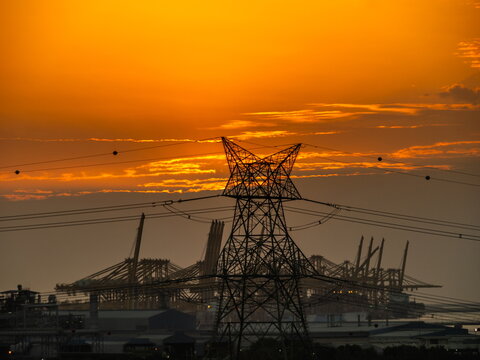 View of a high-voltage power pylon silhouetted against a vibrant orange sunset with industrial port cranes in the background Dubai, Dubai, United Arab Emirates.