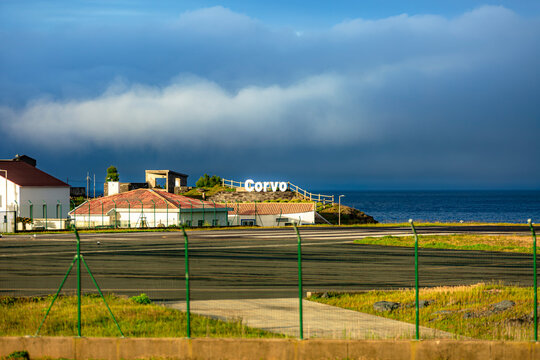 Corvo, Azores, Portugal Small coastal village named Corvo under dramatic clouds. Houses with red-tiled roofs line the shore. Green fence and pathway lead toward the ocean. Blue sea meets sky with stor