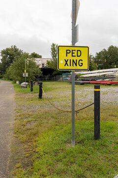 View of a yellow pedestrian crossing sign standing on a metal post near a chain barrier with rowing shells stored in the background in Henrico, Virginia, United States.