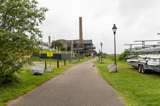 View of a paved path along the riverbank with a rack of rowing shells, a tall brick smokestack, and a modern industrial building in Henrico, Virginia, United States.