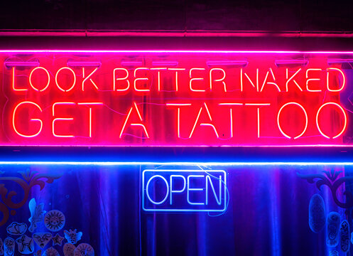View of a neon sign with the text Look Better Naked Get A Tattoo and an Open sign displayed in a tattoo studio window in Portugal.
