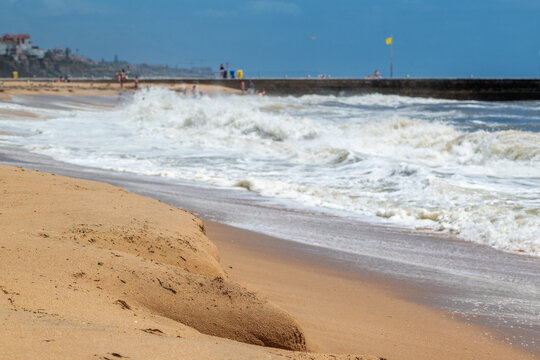 View of a sandy beach showing signs of coastal erosion with crashing waves, foam, and a distant pier under a blue sky in Cascais, Lisbon, Portugal.