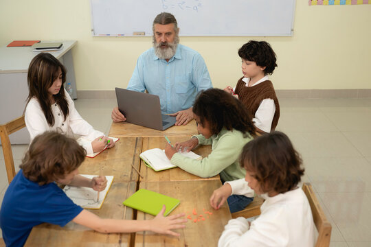 Senior male teacher with a beard teaching math to a diverse group of elementary students sitting around wooden desks in a bright classroom. Concept of inclusive education and collaborative learning.