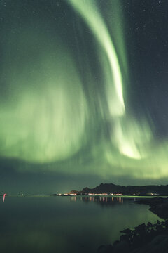 View of vibrant aurora borealis illuminating the night sky above the dark mountains and calm waters of the Lofoten archipelago, Nordland, Norway.