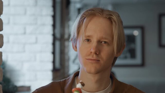 Close up of young man holding and examining food on skewer focusing intently in indoor setting with soft lighting. Background features white brick wall framed pictures creating calm