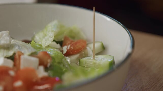 Close up of fresh salad ingredients including lettuce cucumber slices cherry tomatoes and feta cheese held together by toothpick in white bowl on wooden table. Salad components like leafy