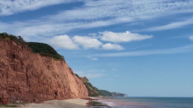 Sidmouth Devon England UK. 14.08.2026. Video. Expansive view of rugged, layered red sandstone cliffs plunging into the sea, with a sandy beach and a horizon stretching to distant headland