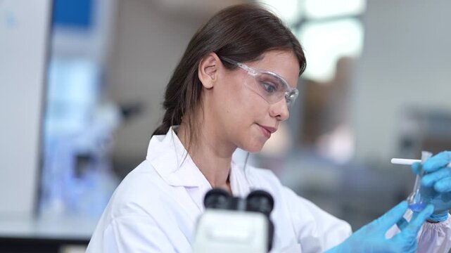 Two women scientists in white lab coats, hairnets and clear safety goggles at a brightly lit laboratory workbench, with focus pulling from the distant bench worker to the colleague in front.