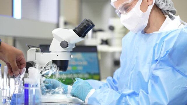 Two female research scientists in white coats, protective eyewear and sterile hairnets at a well-equipped science laboratory bench, with a second hand delivering a frozen sample she then.