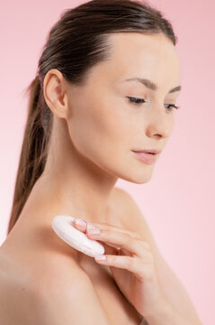 Young woman applying powder to her shoulder with a puff on a pink background