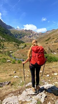 woman hiker with red backpack walking on mountain trail through green valley with trekking poles- Ordesa and Monte Perdido National Park, Spanish Pyrenees