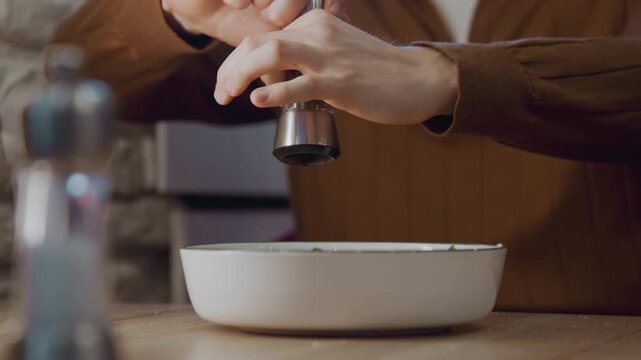 Close up of person in brown clothing seasoning food with pepper into white bowl on wooden table. Pepper grinder and salt shaker visible background includes stone wall