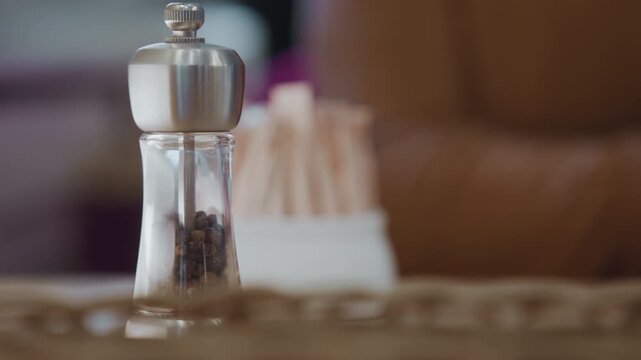 Close up of hand reaching for salt and pepper shakers on table. Hand grasps silver top adjusts position near glass containers filled with black peppercorns and white granulated salt. Soft