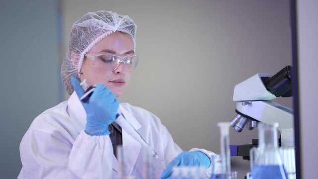A woman researcher in a white coat, protective eyewear and a sterile hairnet raising a micropipette to check the tip then lowering it into a tube in a bright modern life science laboratory.