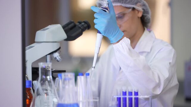 A woman researcher in a white lab coat, hairnet and clear safety goggles dipping the pipette to the rack, lifting it high, then bringing it down again at a brightly lit laboratory workbench.