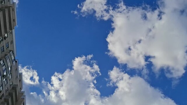 Low angle view of white clouds moving fast across the blue sky over a residential building outdoors. Urban weather conditions, daytime timelapse background, city environment concept.