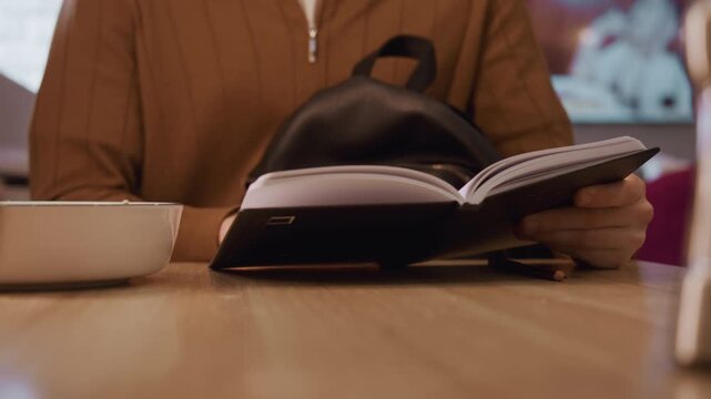 Close up of person flipping through notebook pages at table with pencil and bowl nearby. Hands interact with open book revealing text details. Warm indoor setting with soft lighting creates