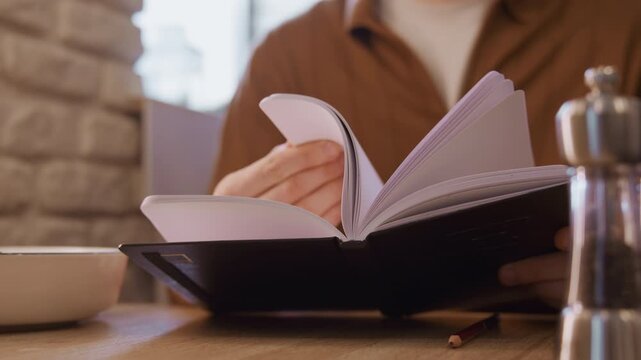 Close up of person flipping through blank pages in open notebook on wooden table. pepper grinder and bowl are visible nearby with soft natural light illuminating scene indoors