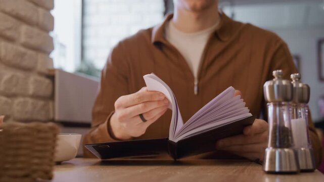 Close up of man flipping through pages of open notebook at wooden table. scene includes visible objects like bowls pepper shakers and soft indoor lighting with natural light from windows