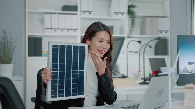 Professional woman presenting a solar panel in a modern office with a laptop and wind turbine model. The scene represents renewable energy, clean technology, sustainability, solar power and the transi