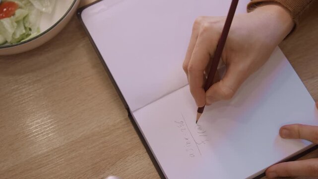 Close up of woman hand writing mathematical equations in pencil on white paper with soft lighting and natural expression. scene shows woman holding open notebook steady while forming precise