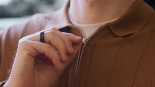 Close up of man unzipping brown ribbed fabric jacket revealing white shirt underneath. Hands with black ring manipulate silver zipper pull tab throughout. Soft lighting highlights texture