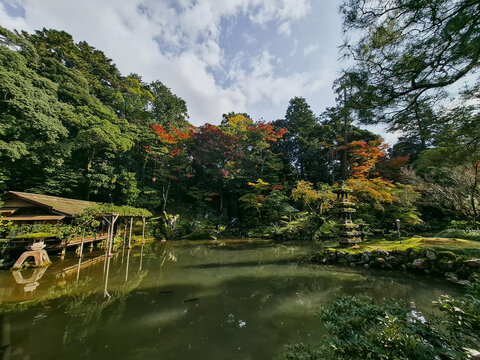 View of a serene pond in Kenrokuen Garden with a traditional wooden teahouse, stone lantern, and vibrant autumn foliage in Kanazawa, Ishikawa, Japan.