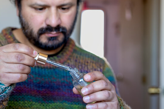Close-up of a bearded man in a colorful knitted sweater using a dropper bottle indoors, suggesting medicine, essential oil, or skincare use