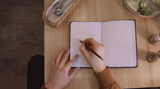 Close up of person writing mathematical equations in open notebook on wooden table. salad with fork woven utensil basket and pepper grinders are visible nearby. Soft natural light