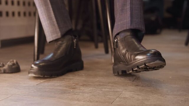 Close up of man feet in black leather boots with thick soles and side zippers slightly shifting position on wooden floor. man wears gray plaid trousers suggesting formal or semi formal