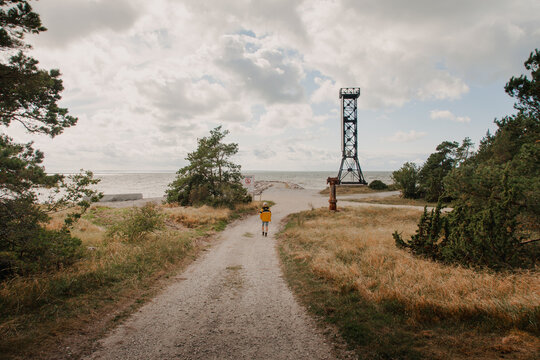 View of a child in a yellow jacket walking along a gravel path towards the Saxby Lighthouse on the coast, with trees and a cloudy sky in the background in Saxby, Laane maakond, Estonia.