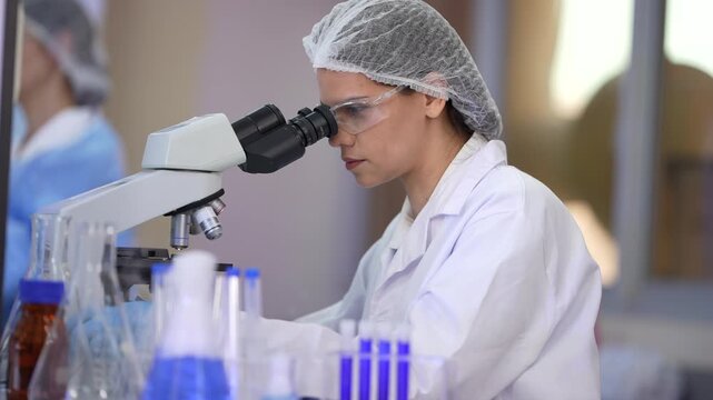Two women scientists wearing blue protective gowns, sterile hairnets and nitrile gloves looking into the eyepieces, glancing up at a colleague, then returning to the slide at a brightly lit.