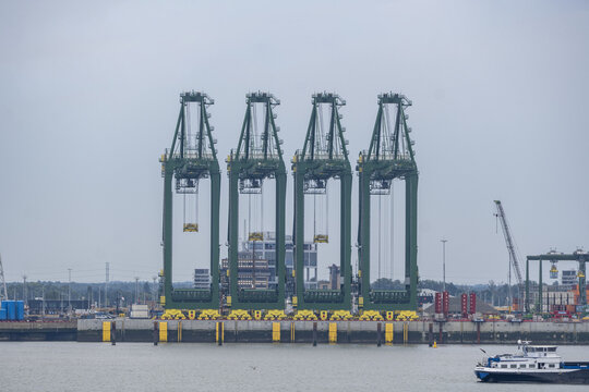 View of Port of Antwerp-Bruges International with four green container cranes along the quay, a barge in the foreground, and industrial infrastructure Antwerpen, Vlaanderen, Belgium.