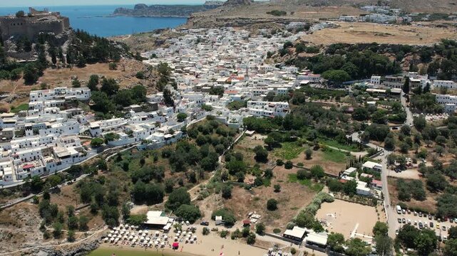 Stunning aerial drone footage flying over the ancient Acropolis of Lindos in Rhodes, Greece. The footage reveals the historic ruins perched above the picturesque whitewashed village of Lindos