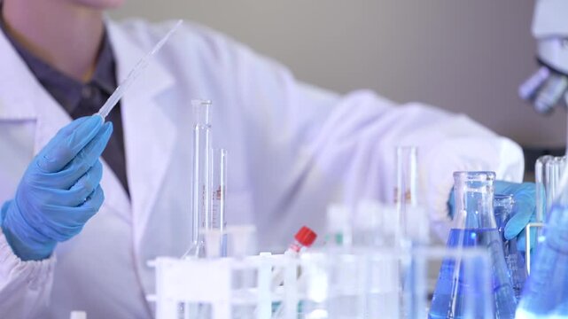 The nitrile-gloved hands of a female lab technician moving a flask aside and reaching a dropper down toward the tube rack at a professional laboratory workstation.