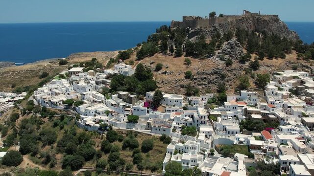 Stunning aerial drone footage flying over the ancient Acropolis of Lindos in Rhodes, Greece. The footage reveals the historic ruins perched above the picturesque whitewashed village of Lindos