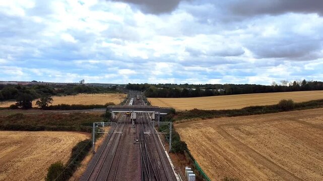 Aerial view of a train traveling along railway tracks through golden wheat fields under a cloudy sky in Finedon, England, United Kingdom.